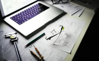 A product design desk with a laptop, technical drawings, and tools